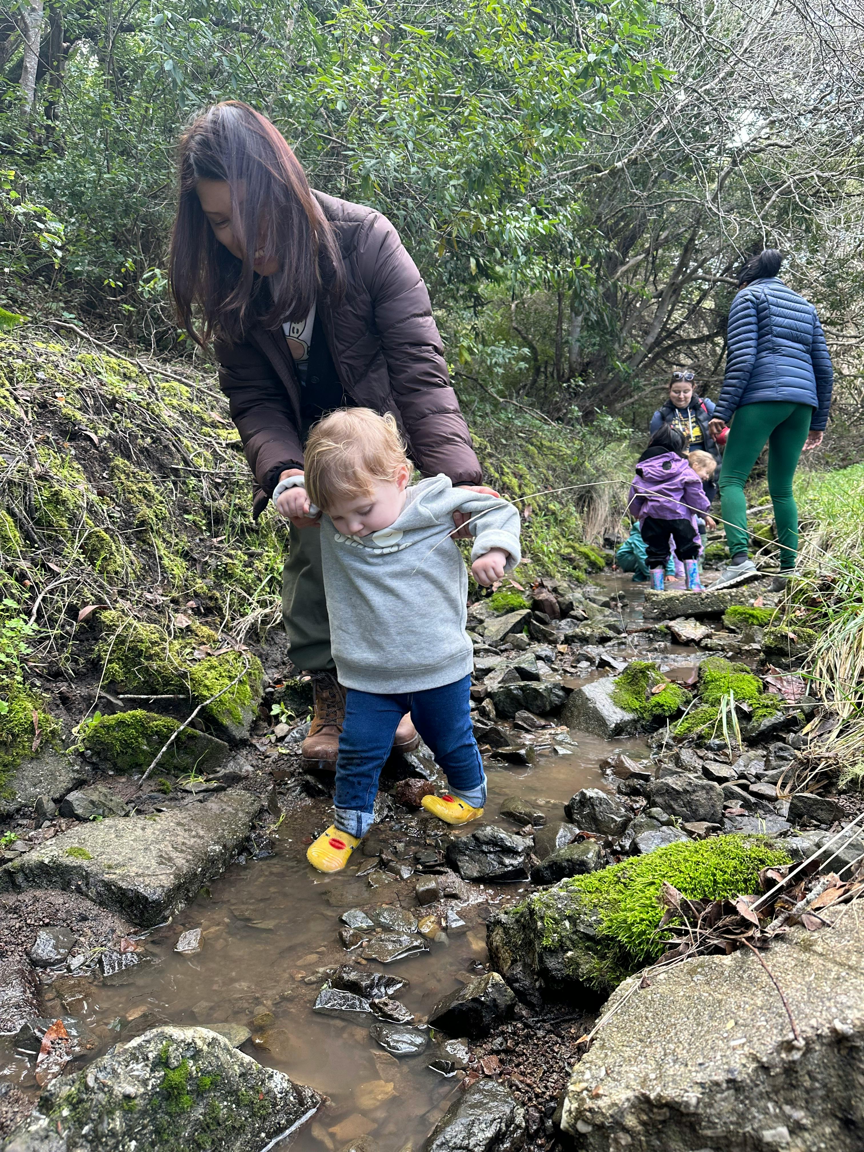 Person helping a child walk through a shallow stream outdoors