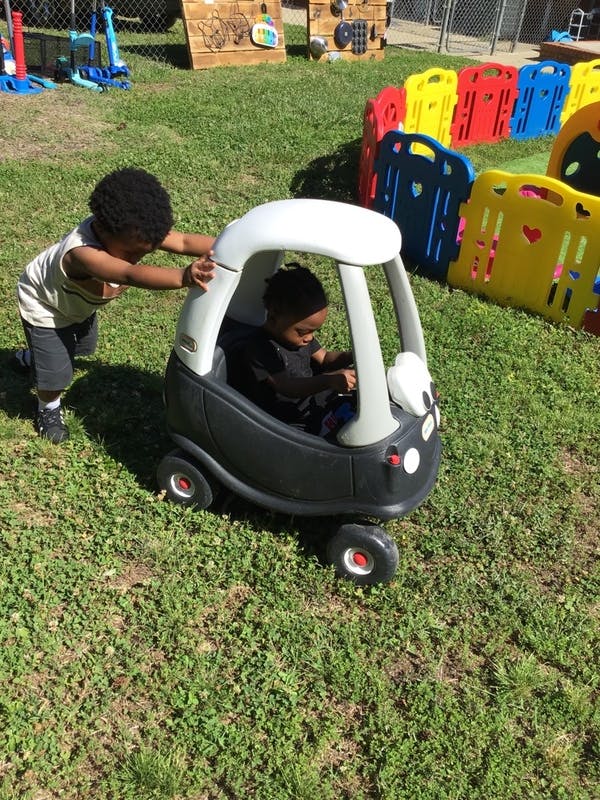 Child pushing another child in a toy car on grass