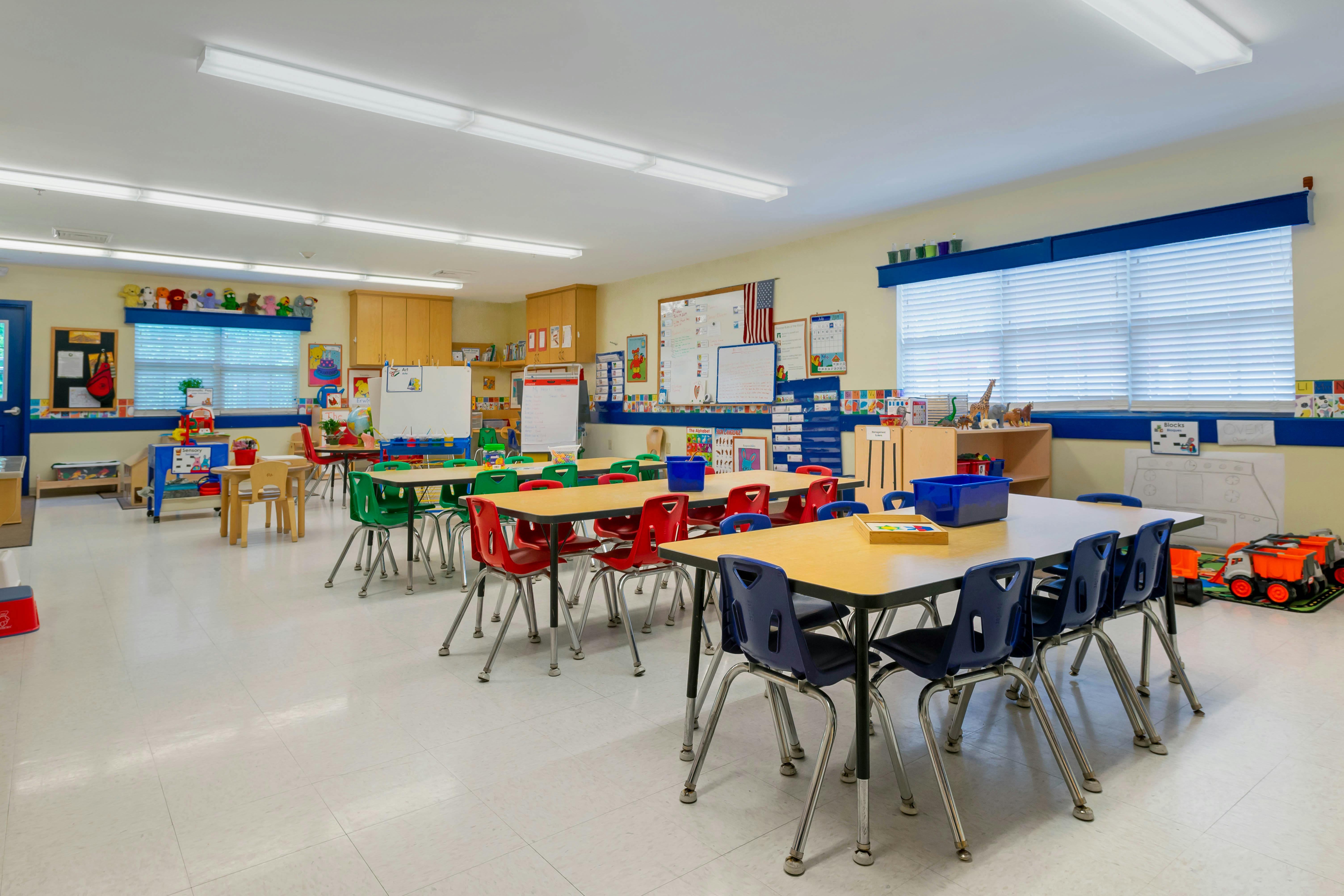 Classroom with multiple tables and chairs, whiteboards, windows, and shelving