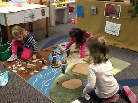 Three children playing with toys on a river-themed rug in a classroom