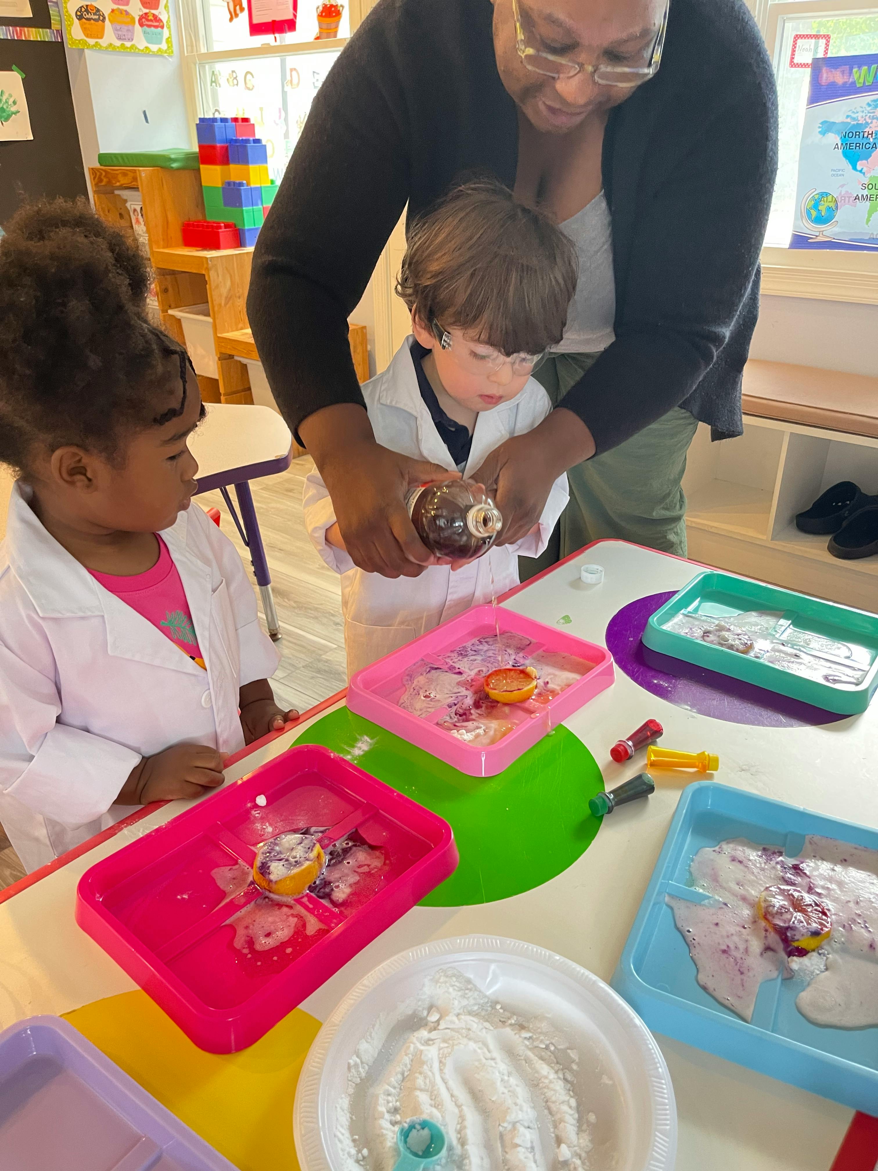 Adult helping a child pour liquid into a tray, another child watching