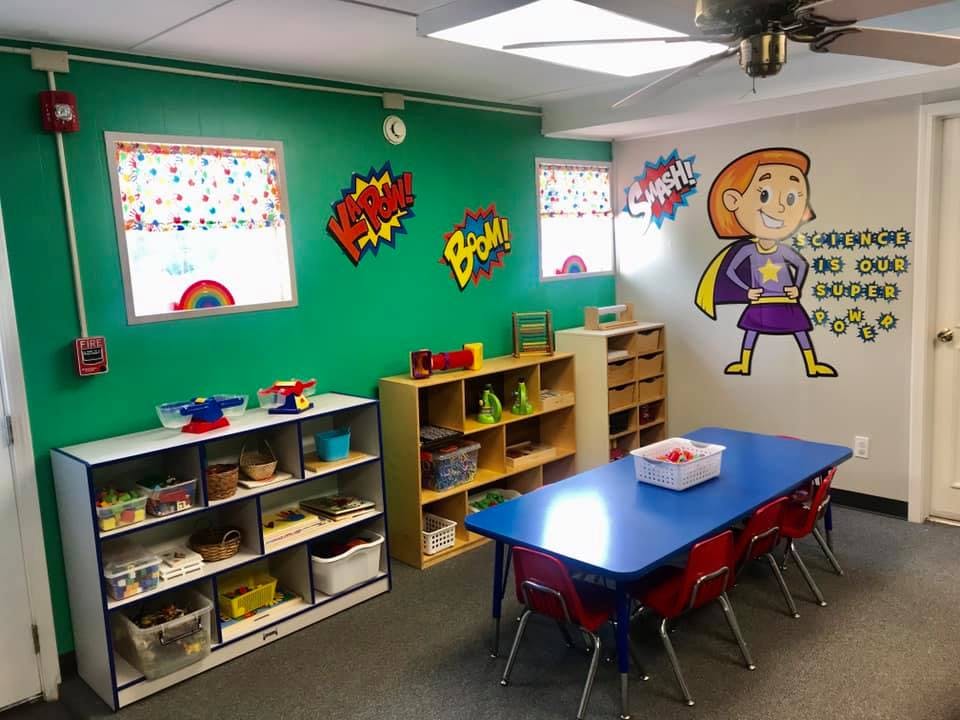 Classroom with green walls, shelves of toys, and a blue table with red chairs