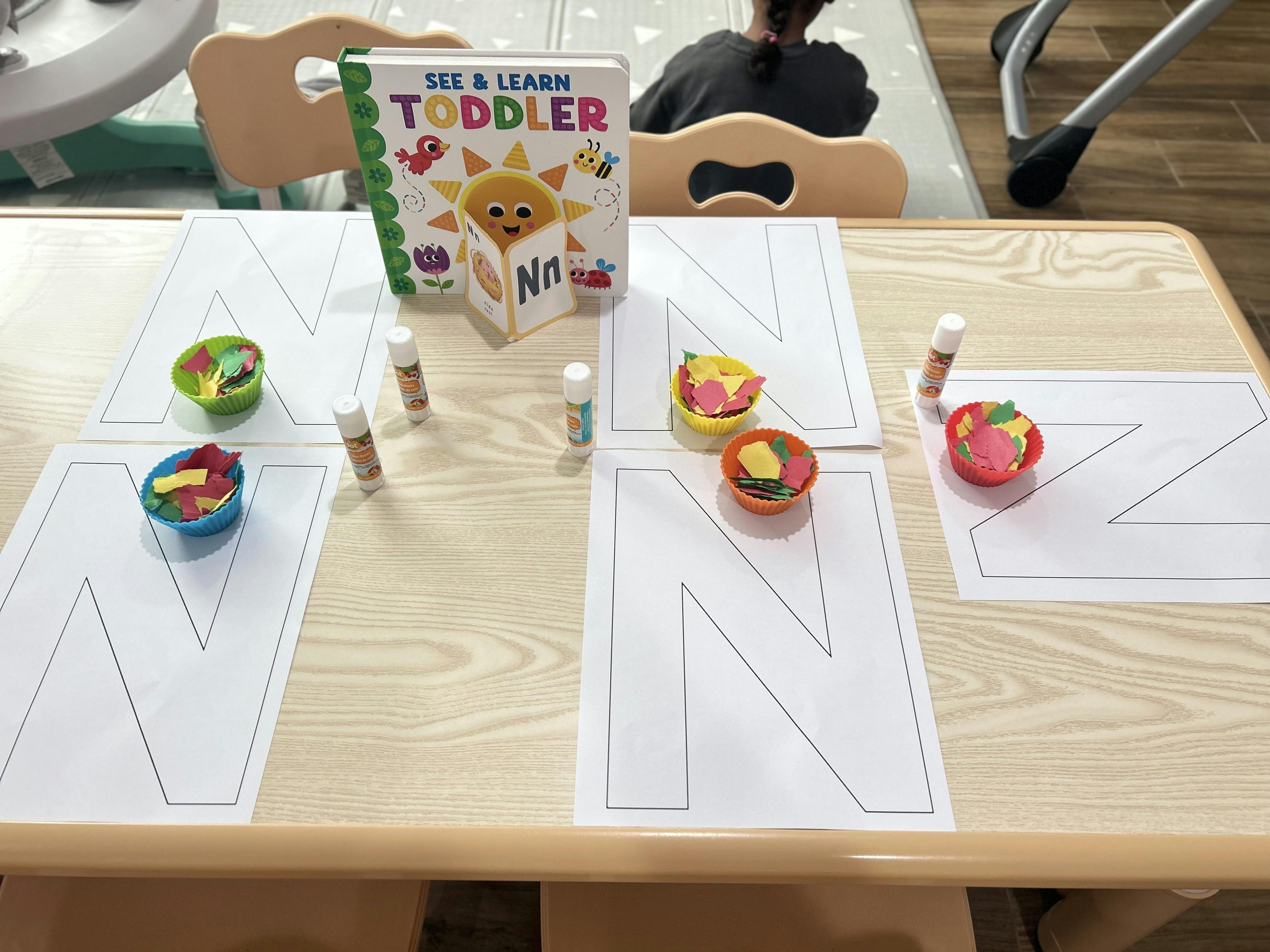 Craft table with letter N outlines, cups of paper, glue sticks, and a toddler book