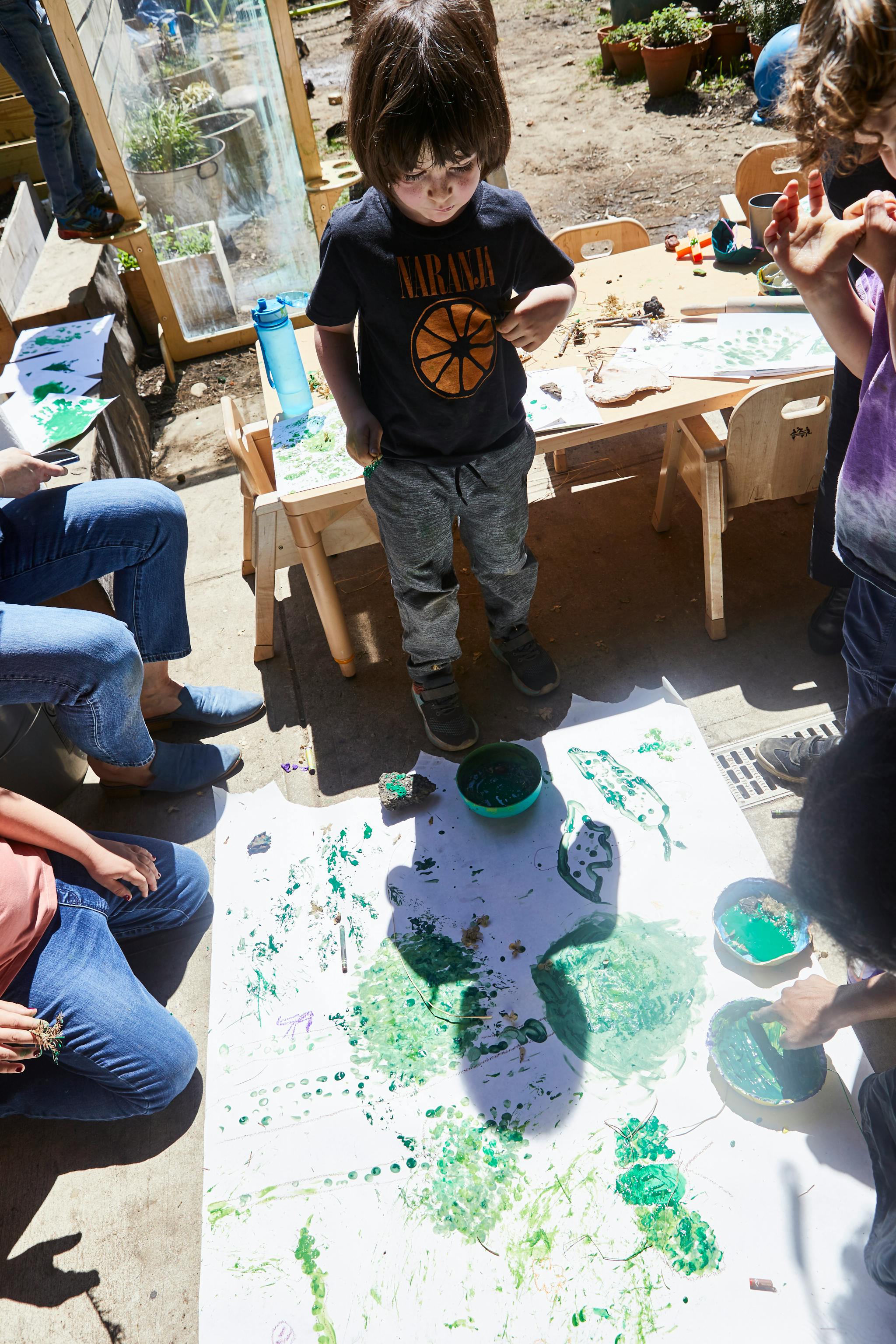 Child standing over a large paper with green paint, other people nearby