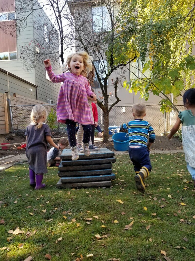 Child jumping on a stack of wooden planks outdoors with other children nearby