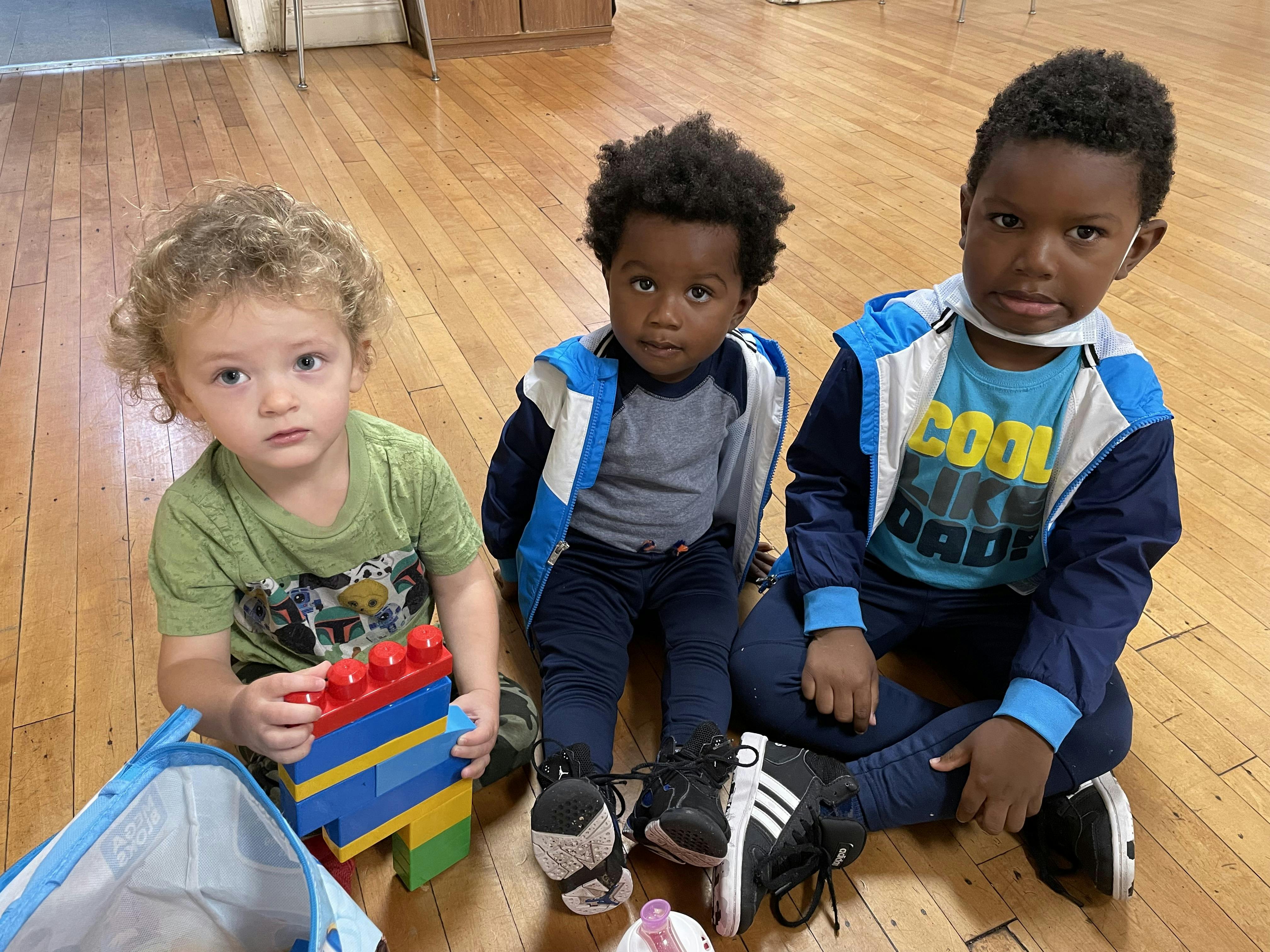 Three children sitting on a wooden floor with toy building blocks