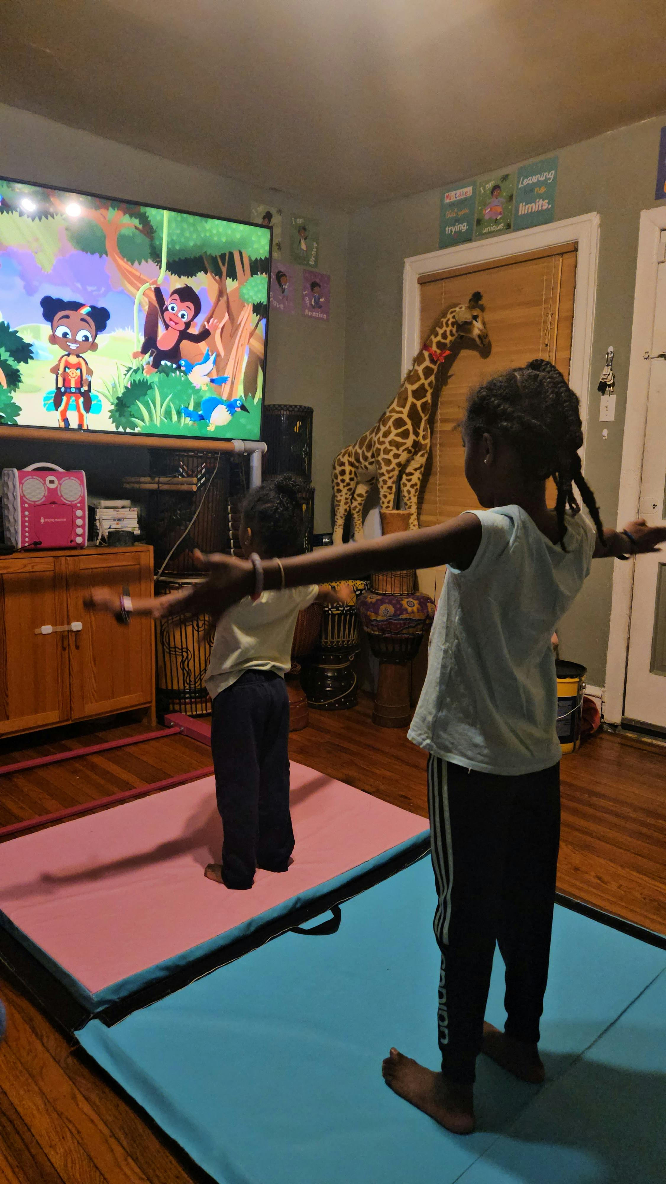 Two children standing on yoga mats with arms outstretched, mimicking a TV