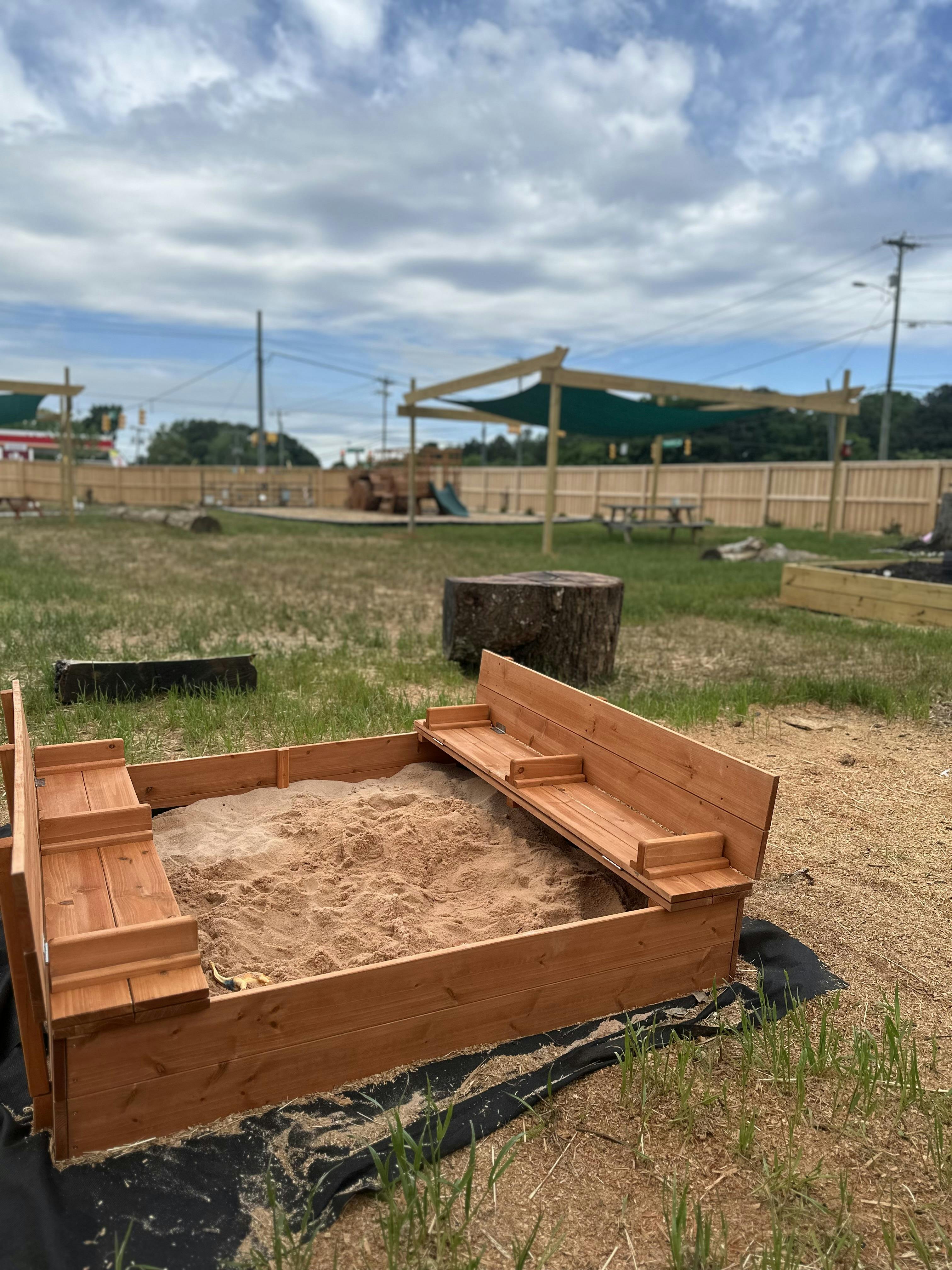 Wooden sandbox filled with sand in a grassy outdoor area with other play structures