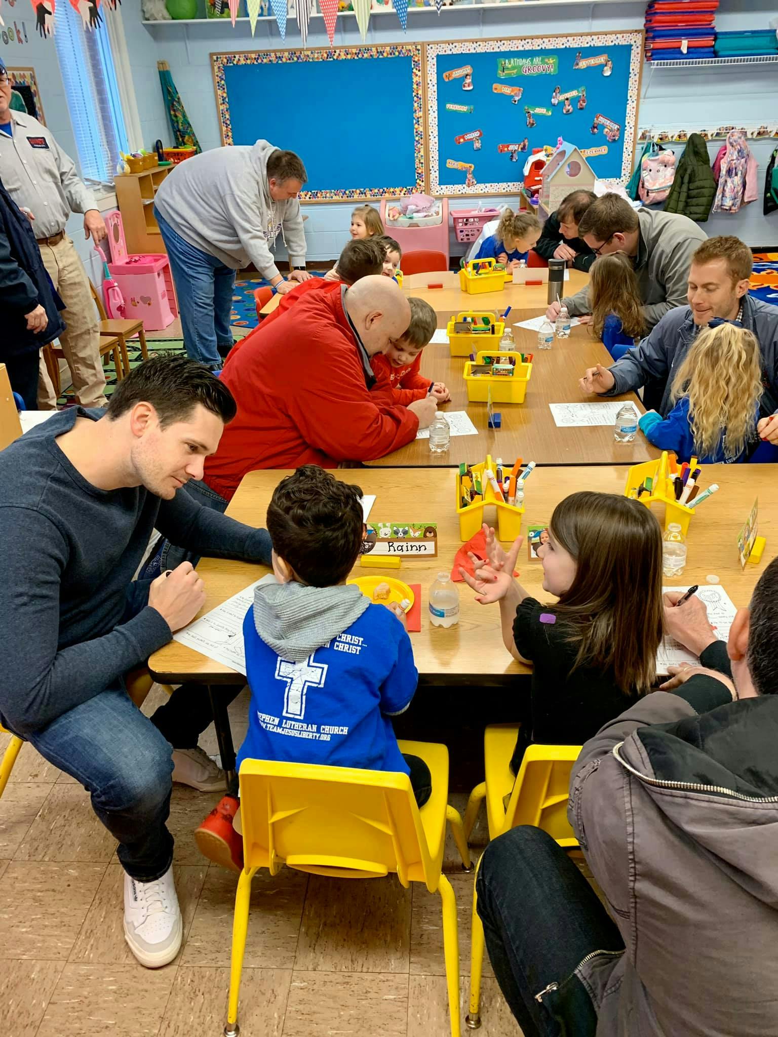 Adults and children sitting at tables in a classroom, engaged in an activity