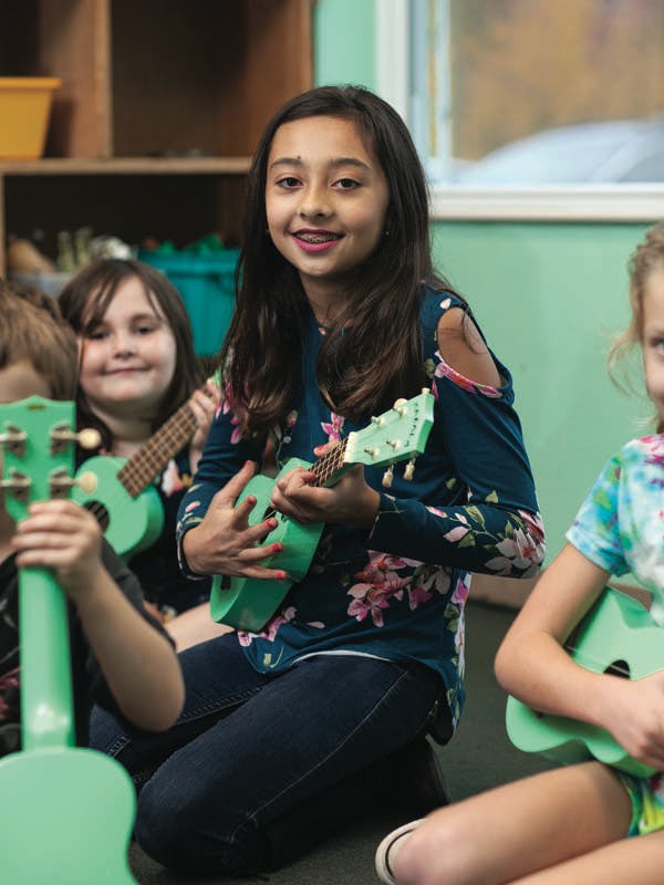 Child playing a green ukulele in a classroom with other children