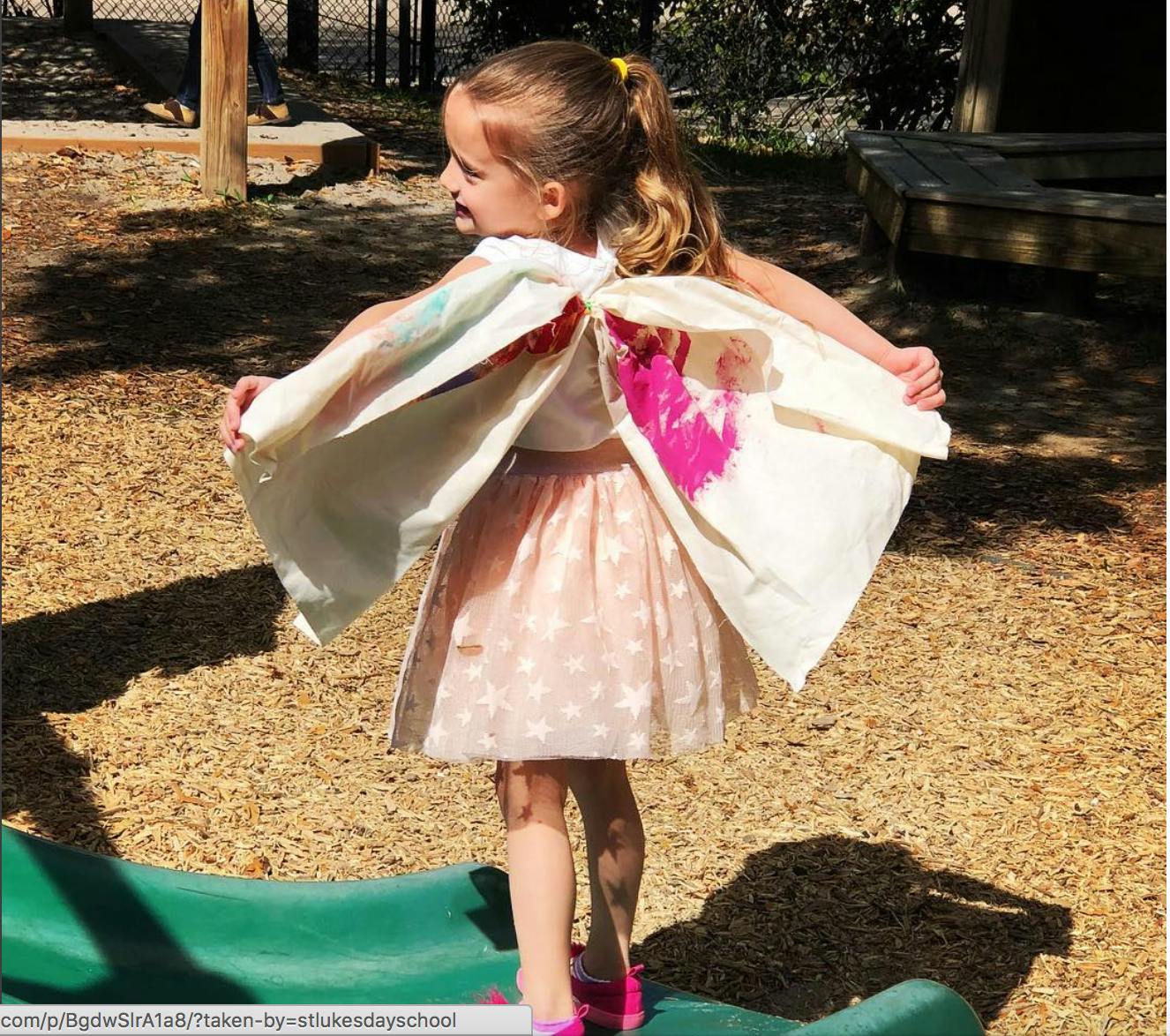 Child in a skirt and cape standing on a green surface outdoors structure