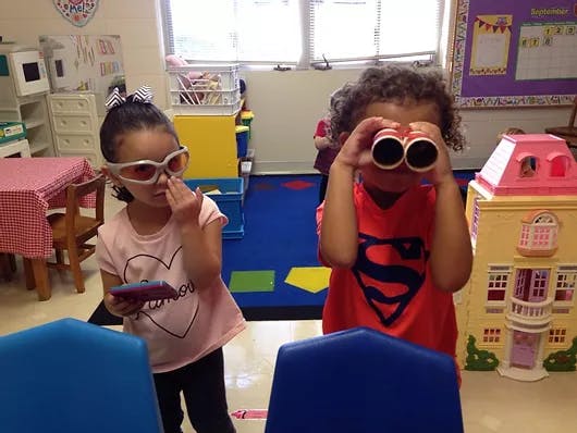 Two children playing in a classroom, one with toy binoculars, one with toy glasses