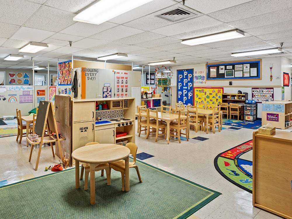 Empty classroom with small tables and chairs, play kitchen, and colorful rugs