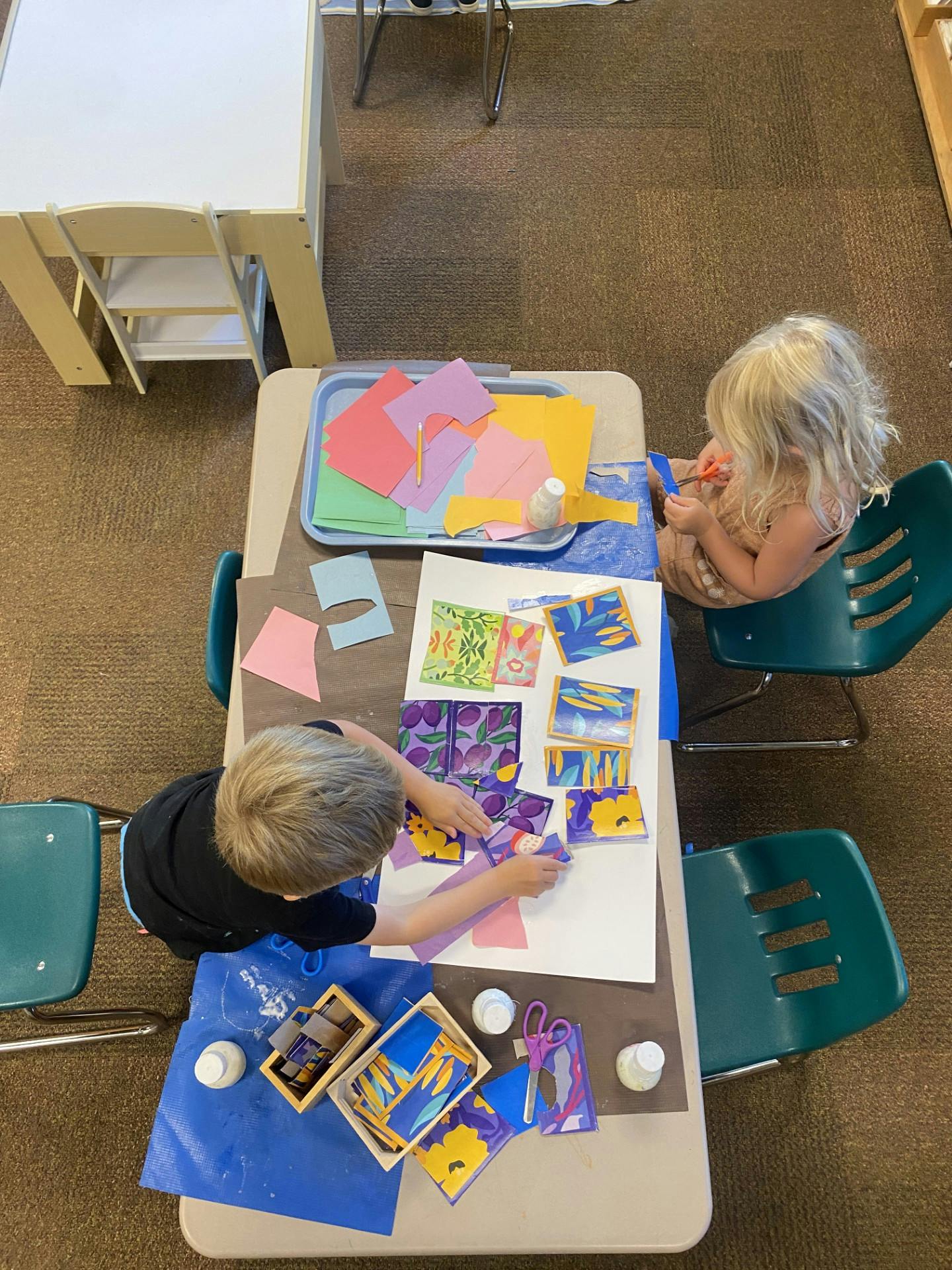 Two children at a table doing crafts with paper, glue, and scissors