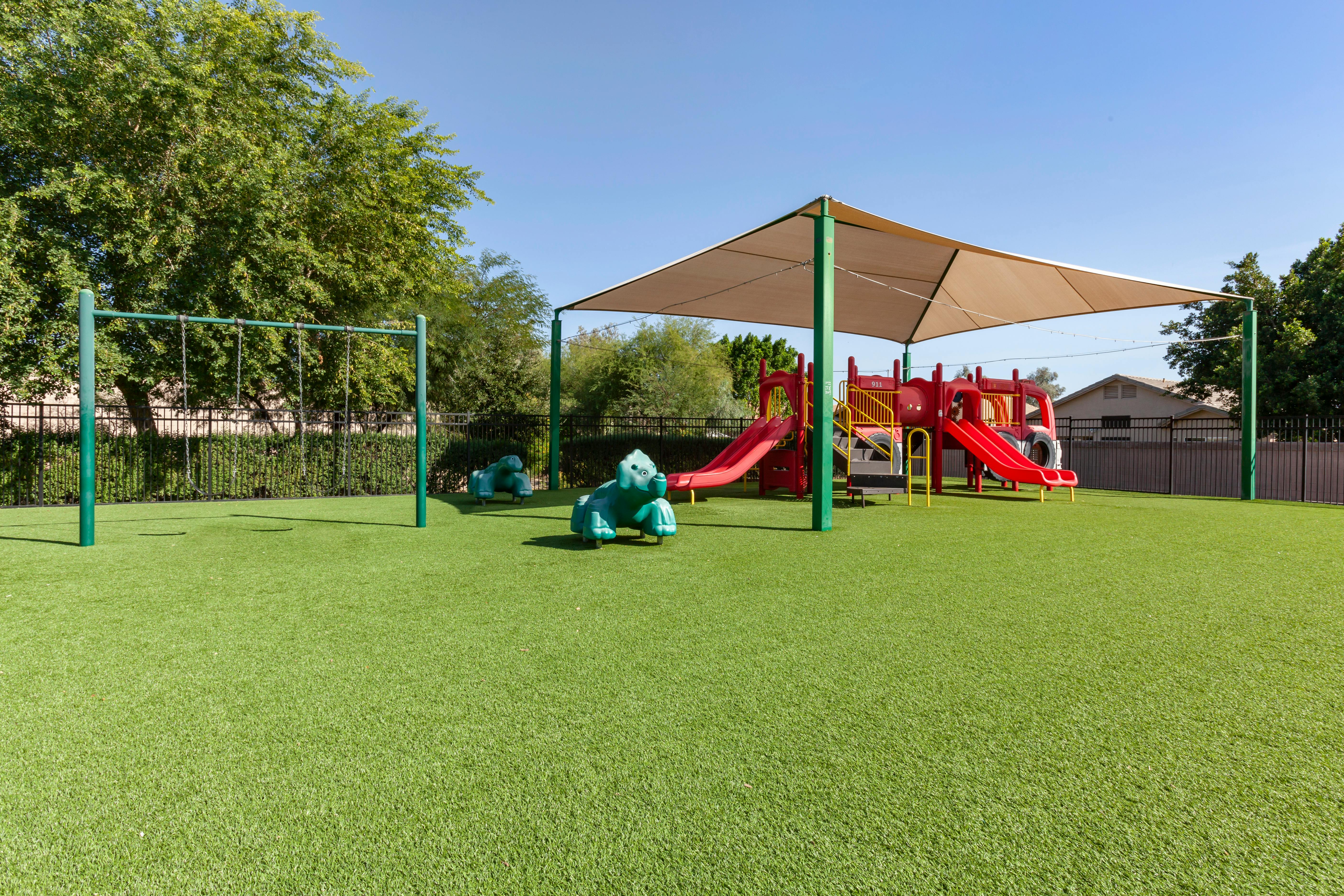 An outdoor playground with swings, red slides, and a tan shade sail
