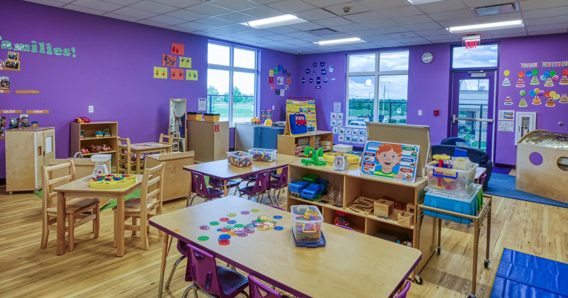 Colorful preschool classroom with tables, chairs, learning materials, and large windows