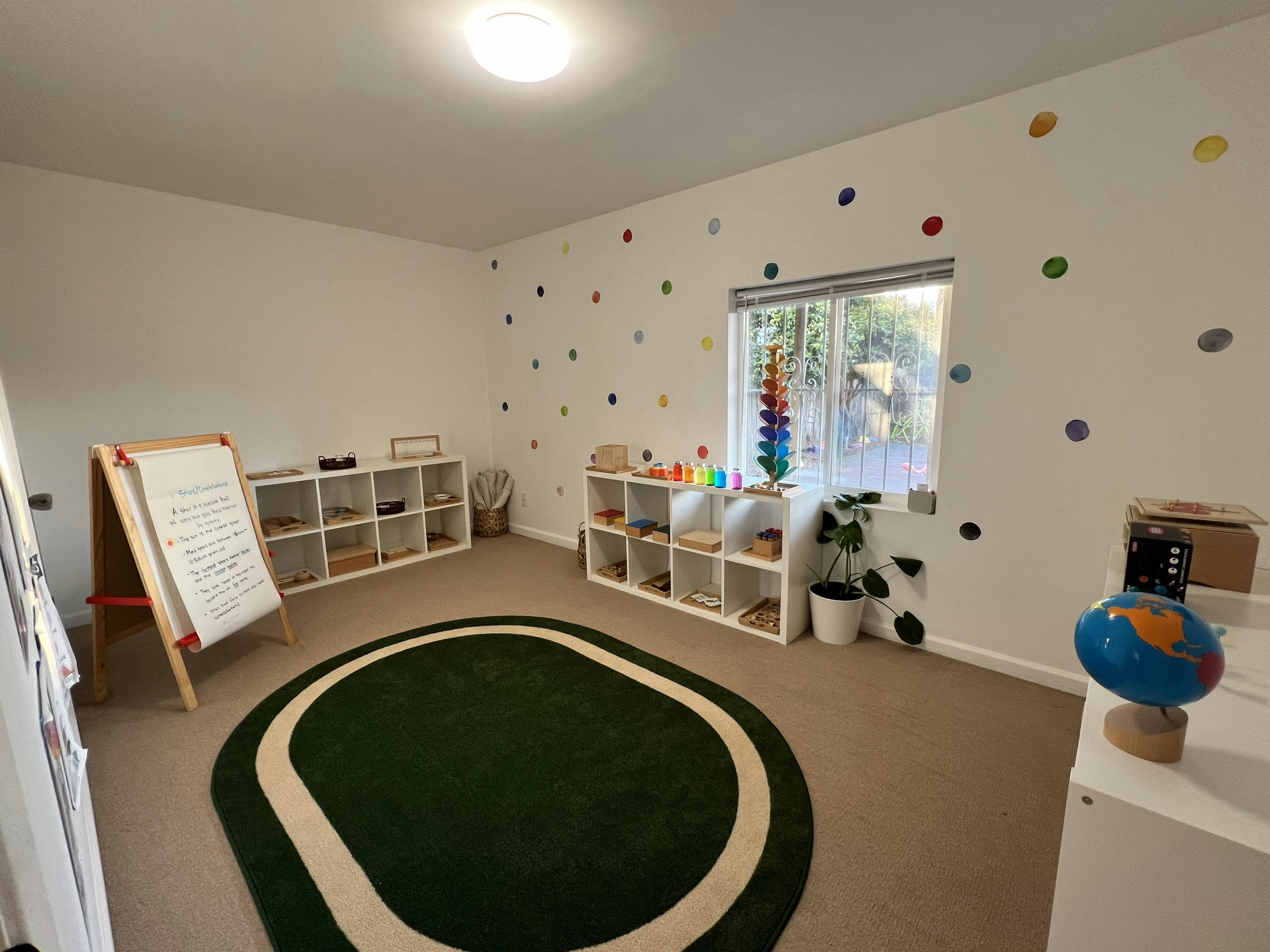 Classroom interior with Montessori-style shelves, a round rug, and easel