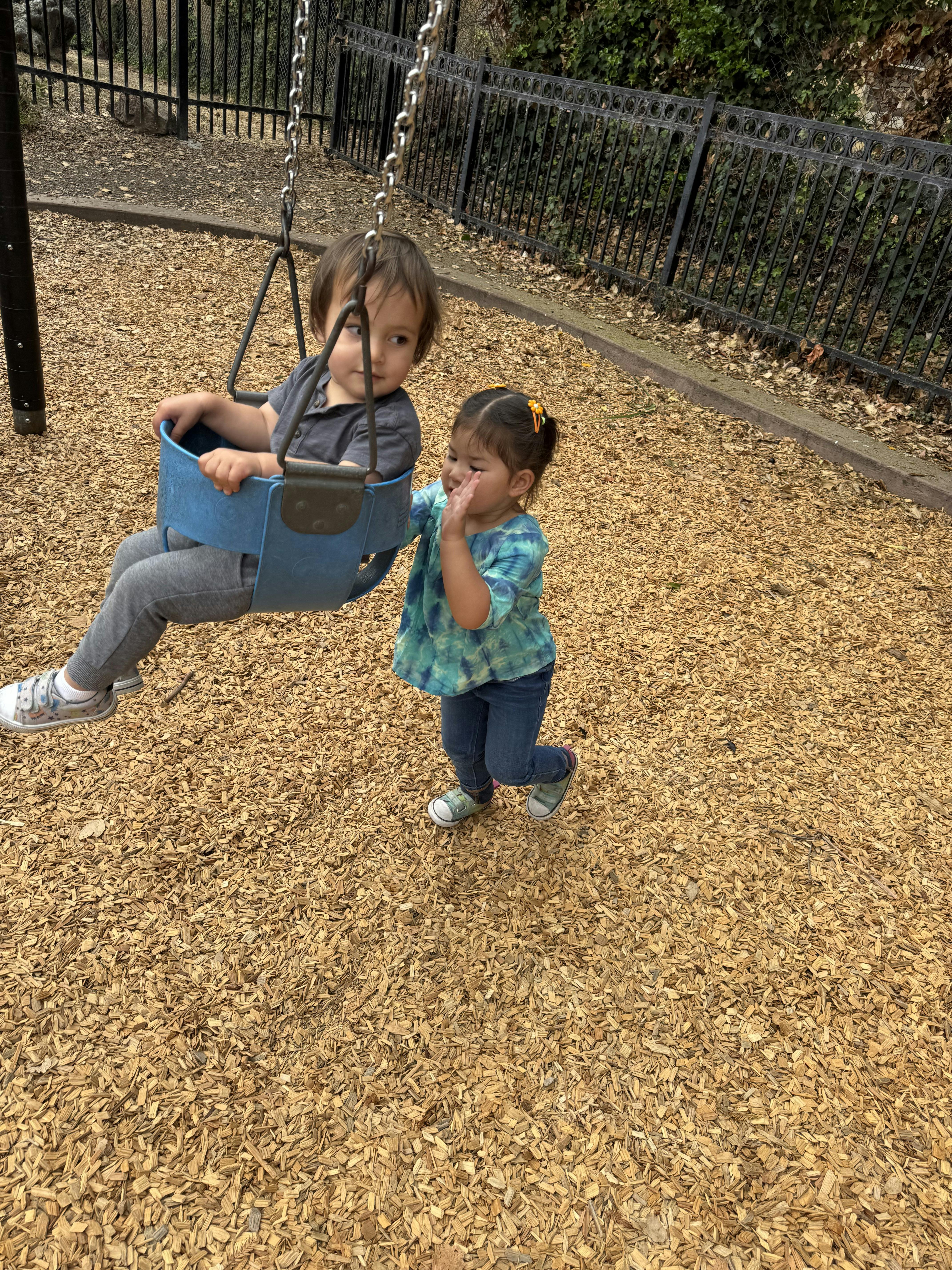 Child in a swing with another child standing nearby on a playground