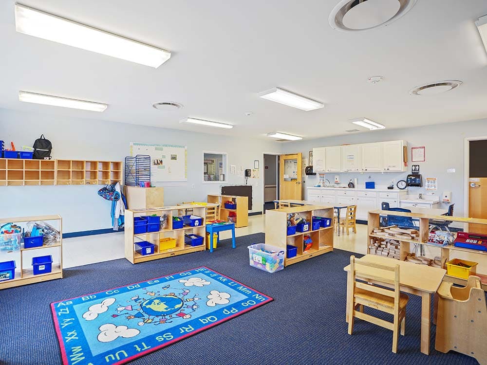 Empty classroom with small wooden tables, chairs, shelving units, and a colorful rug