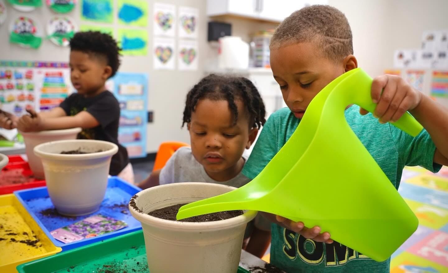 Child pouring soil from a watering can into a pot, with another child watching