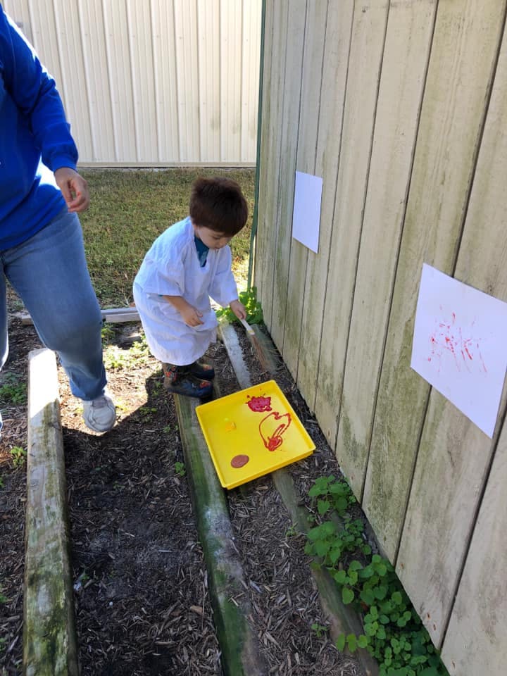 Child painting on paper attached to a wooden wall outdoors