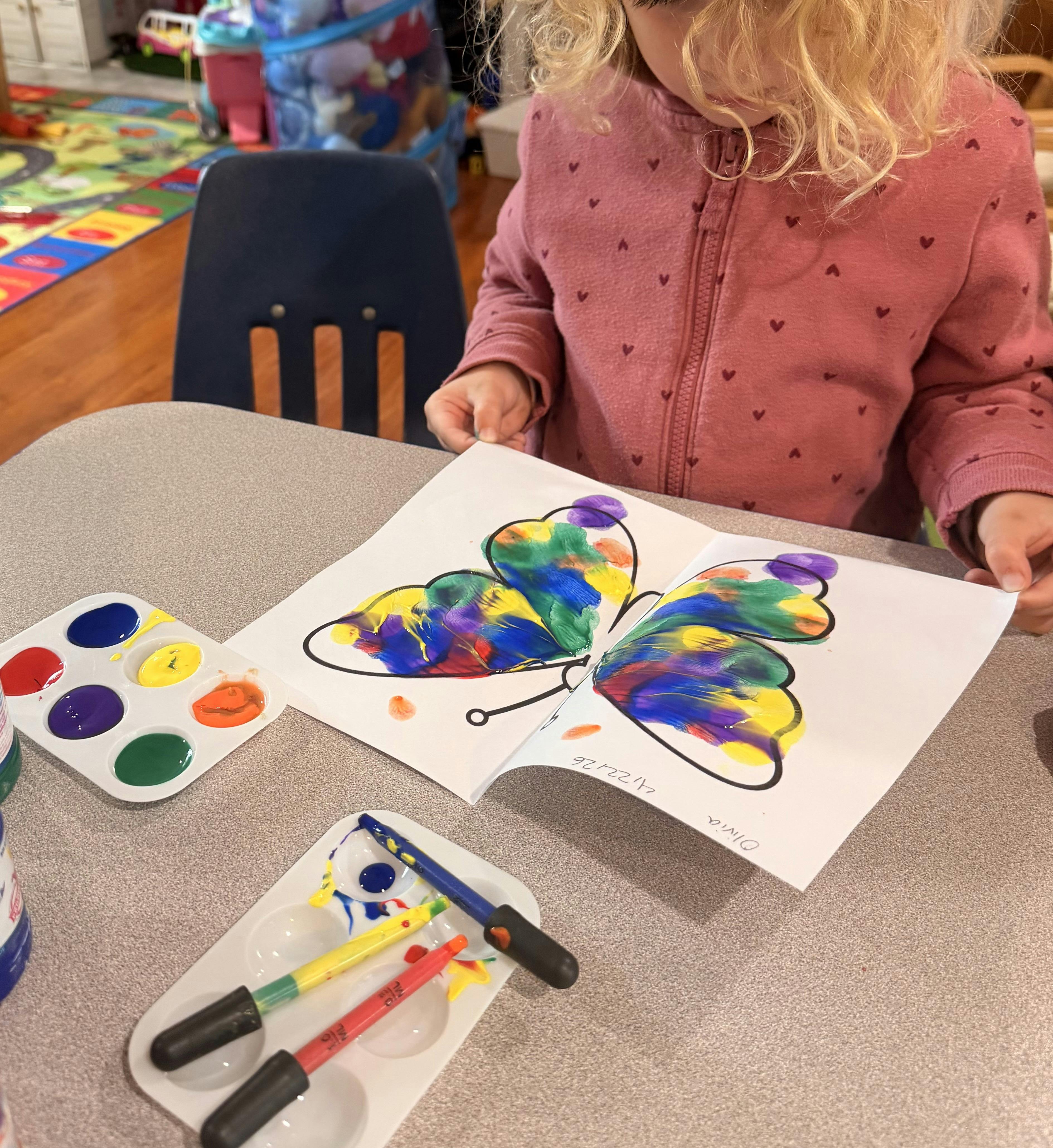 Child in pink top holding paper with a painted butterfly, paint palettes on table