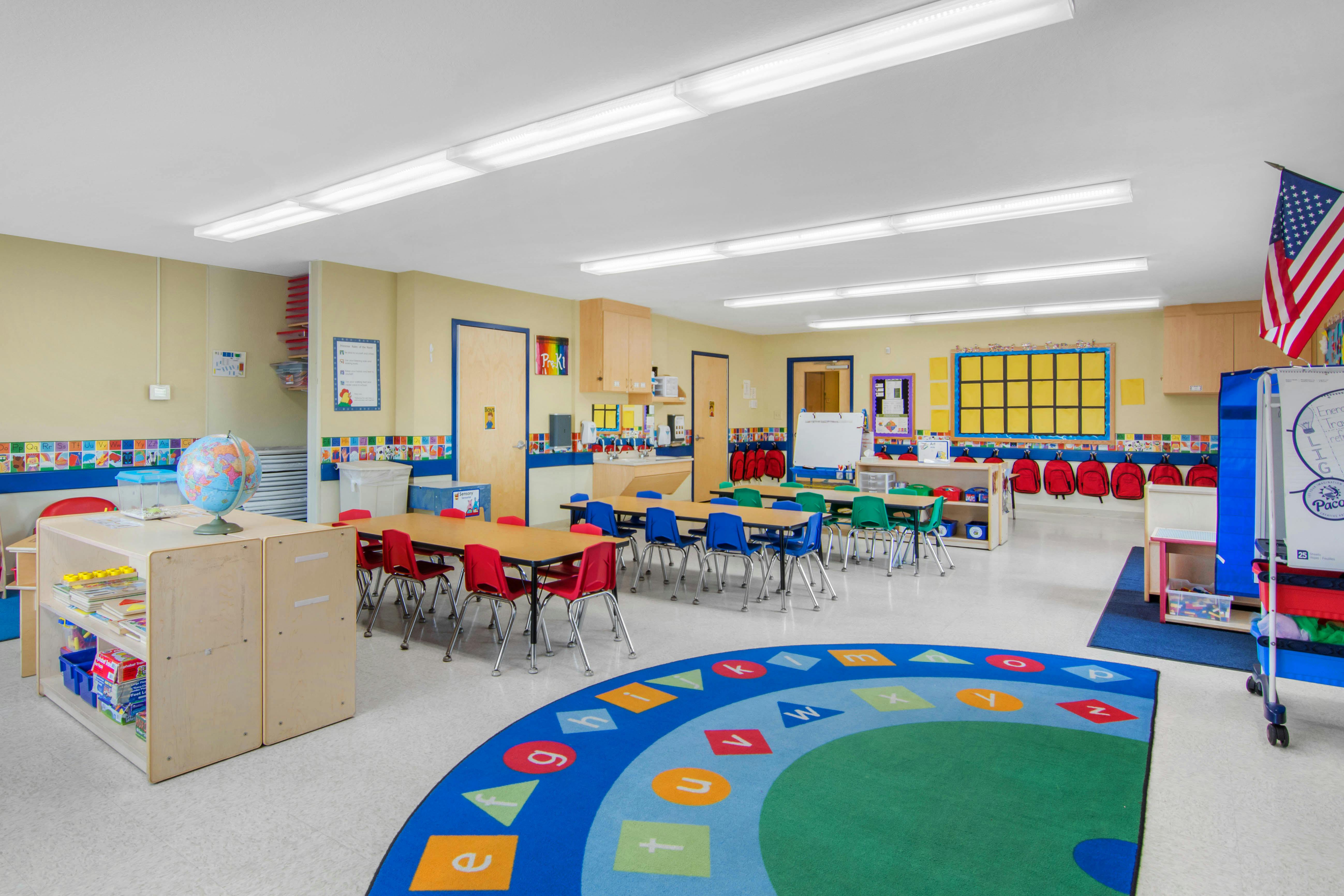 Colorful preschool classroom with small tables and chairs, a round alphabet rug