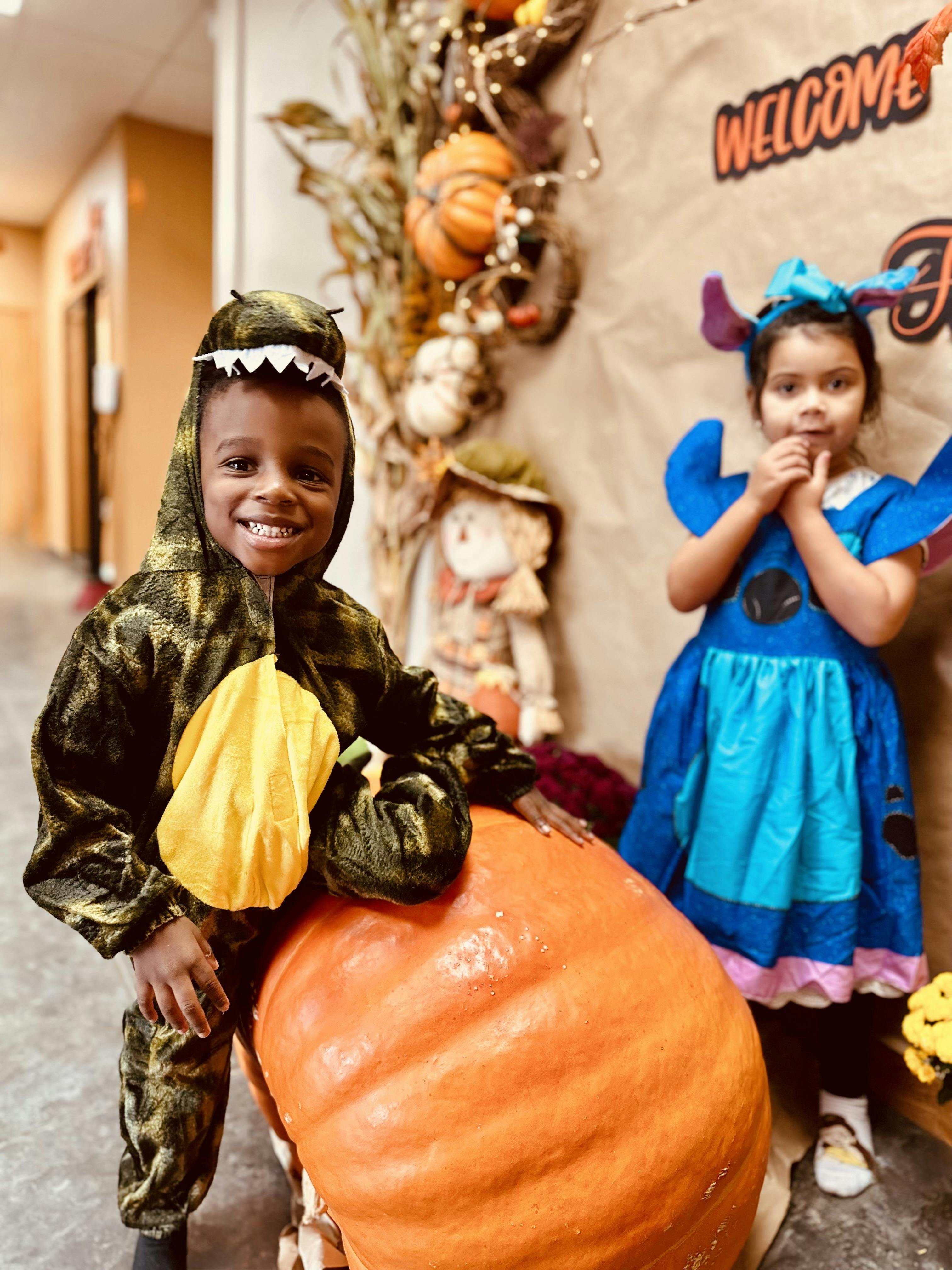 Child in dinosaur costume next to a large pumpkin, another child in blue costume