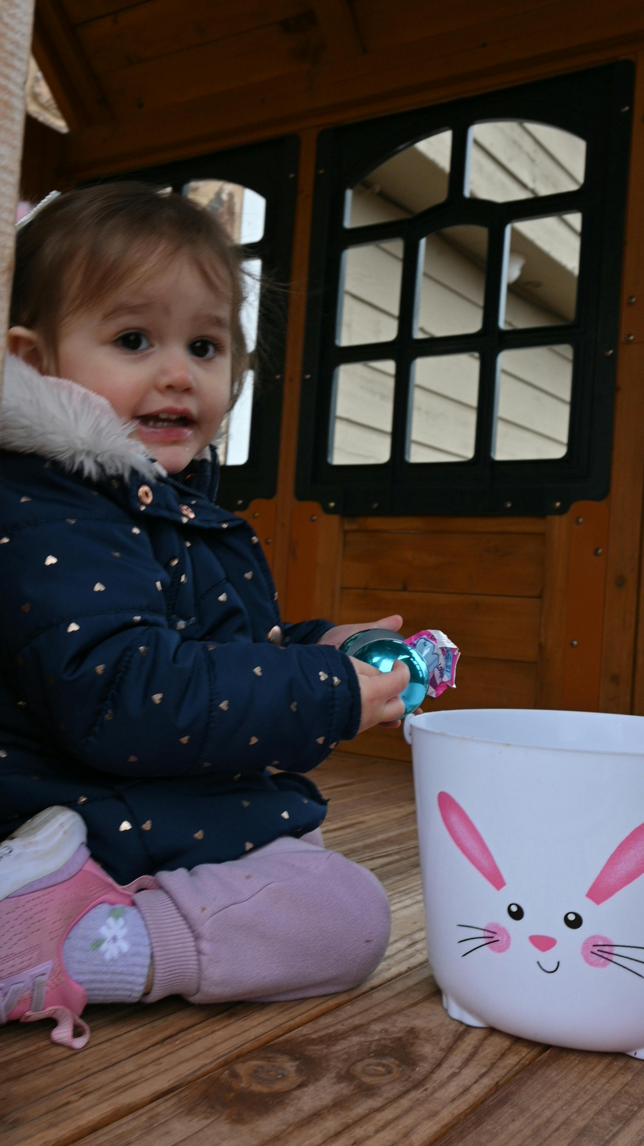 Child in a playhouse holding a blue egg next to a bunny bucket
