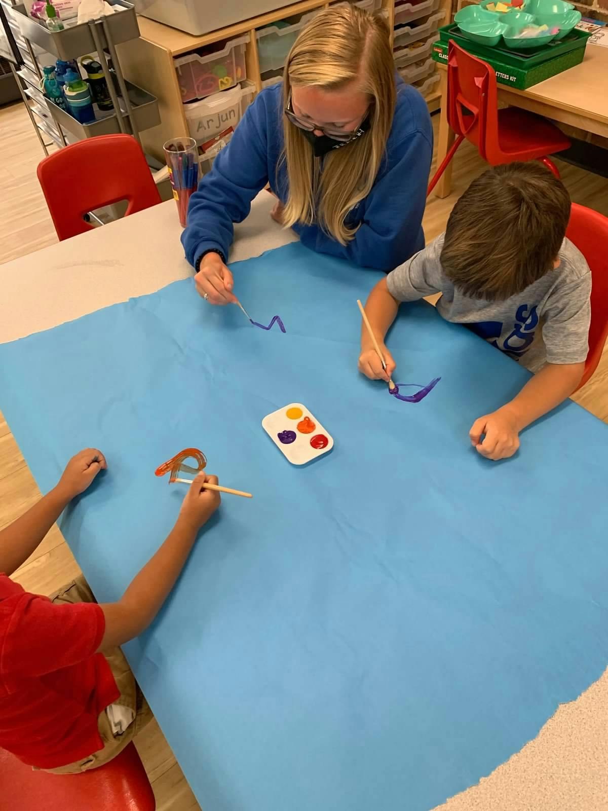 Person and two children painting on blue paper in a classroom