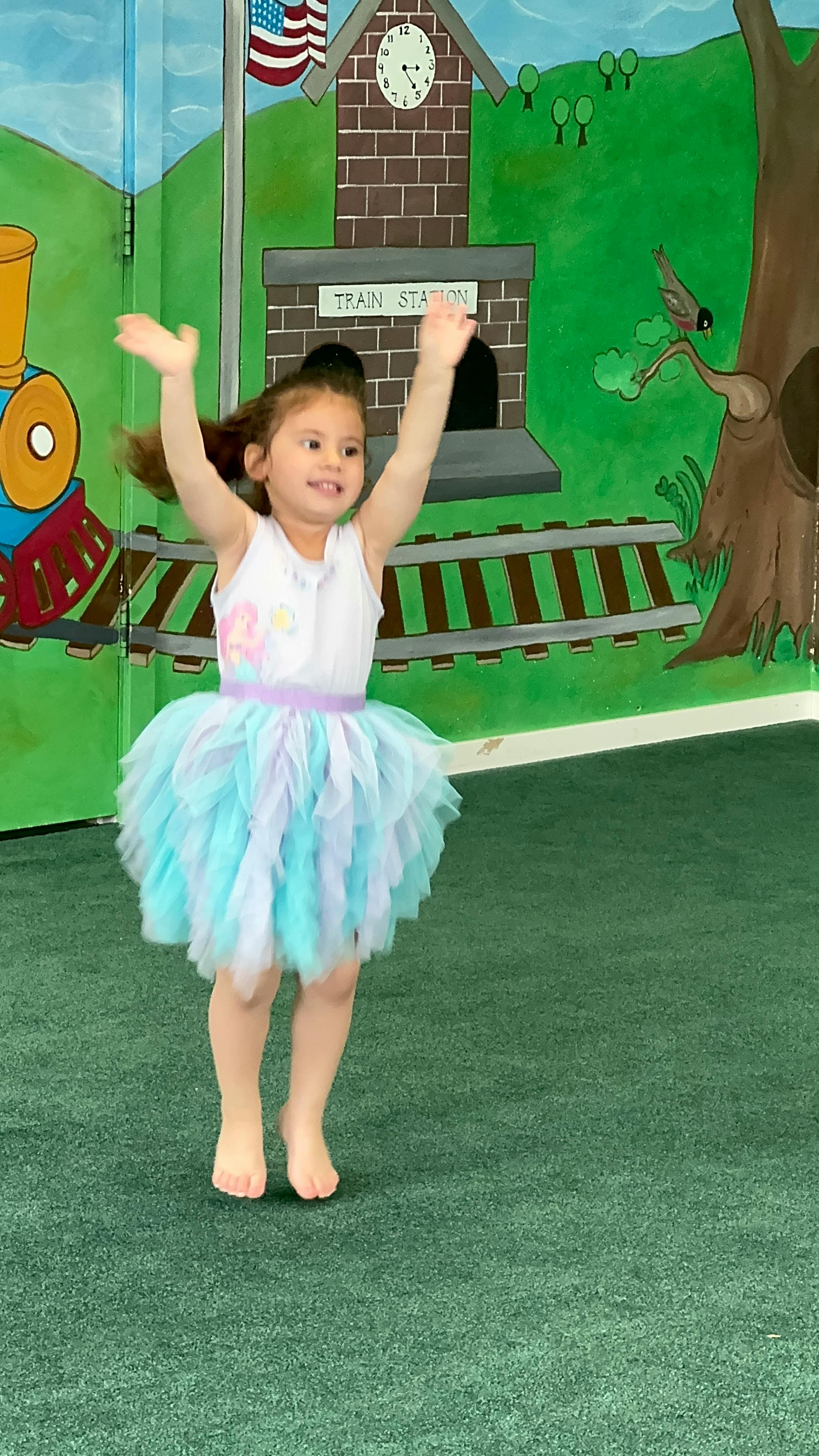 Child in a tutu jumping with arms raised in front of a train station mural