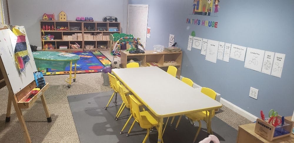 Classroom with a long table and yellow chairs, shelves with toys, and an easel