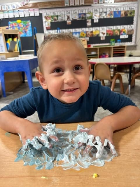 Child with hands in blue and white shaving cream on a table in a classroom