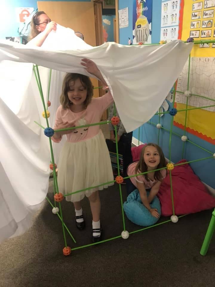 Two children playing in a fort made of green sticks and white sheets