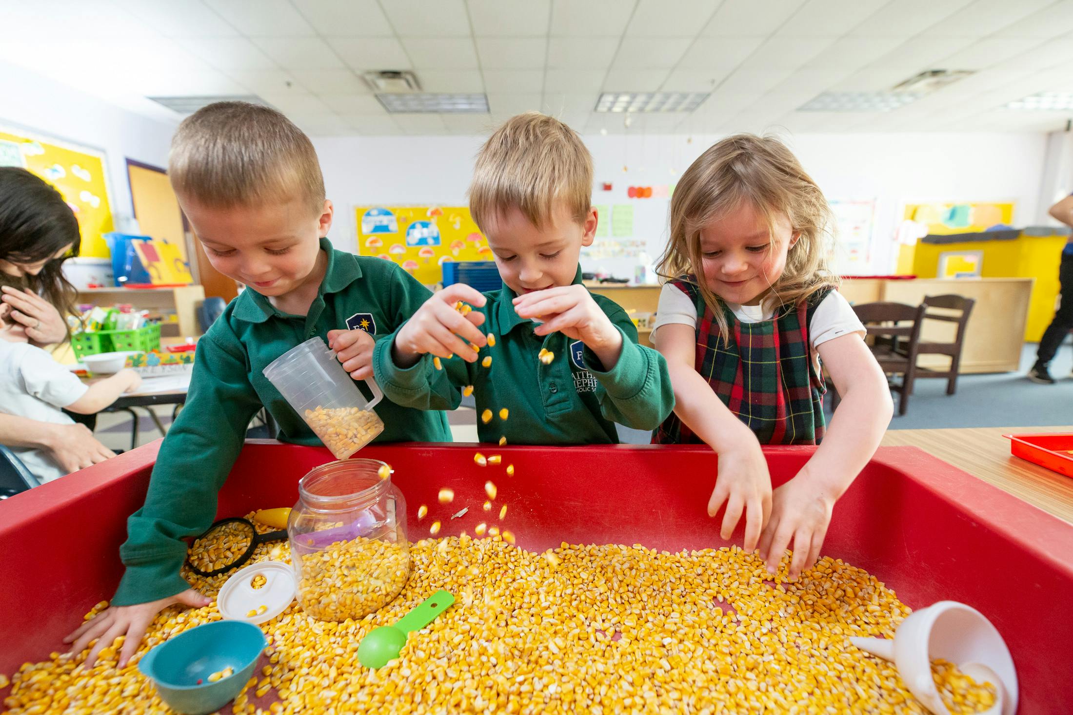 Three children playing with corn kernels in a red bin