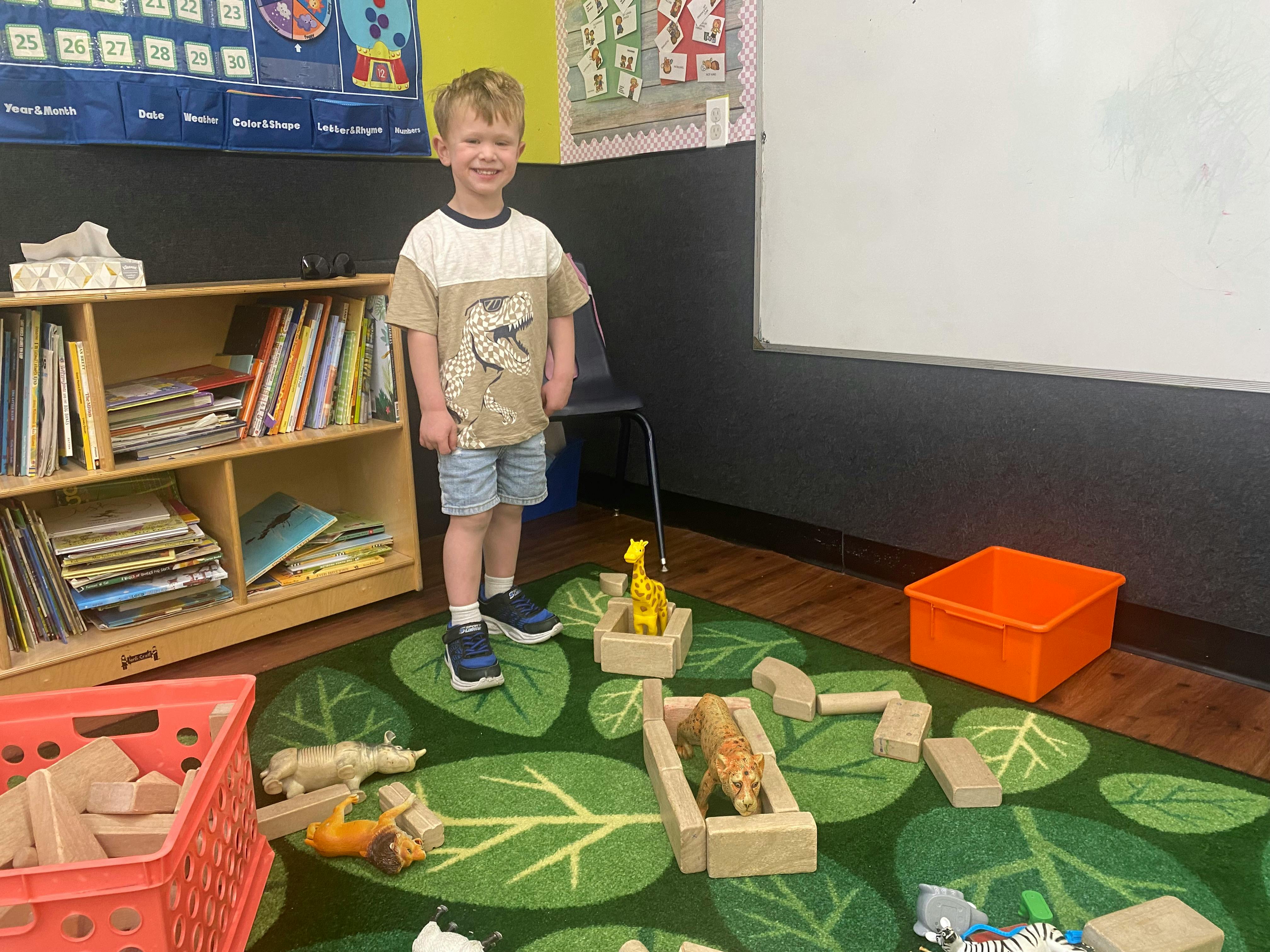 Smiling child standing on a leaf rug surrounded by toys in a classroom