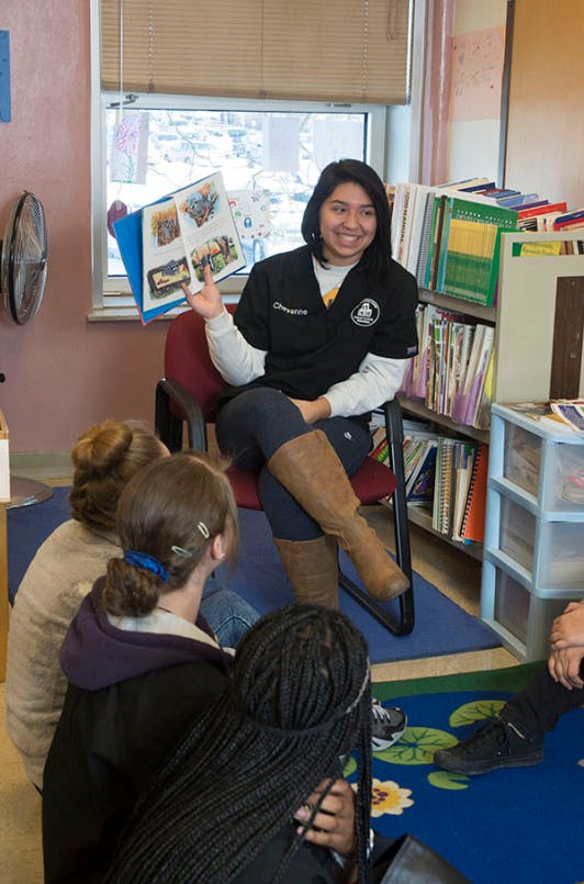 Person sitting in a chair holding an open book and speaking to a group of people