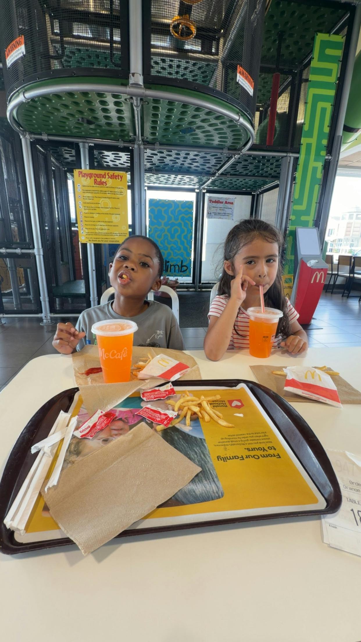 Two children at a table with drinks and fries, playground structure in background