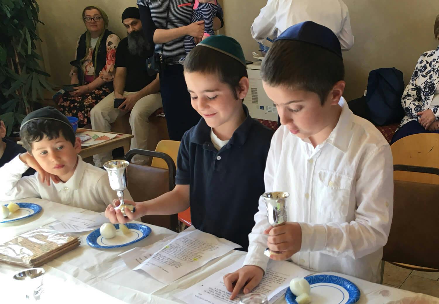 Three children at a table holding kiddush cups, reading from papers