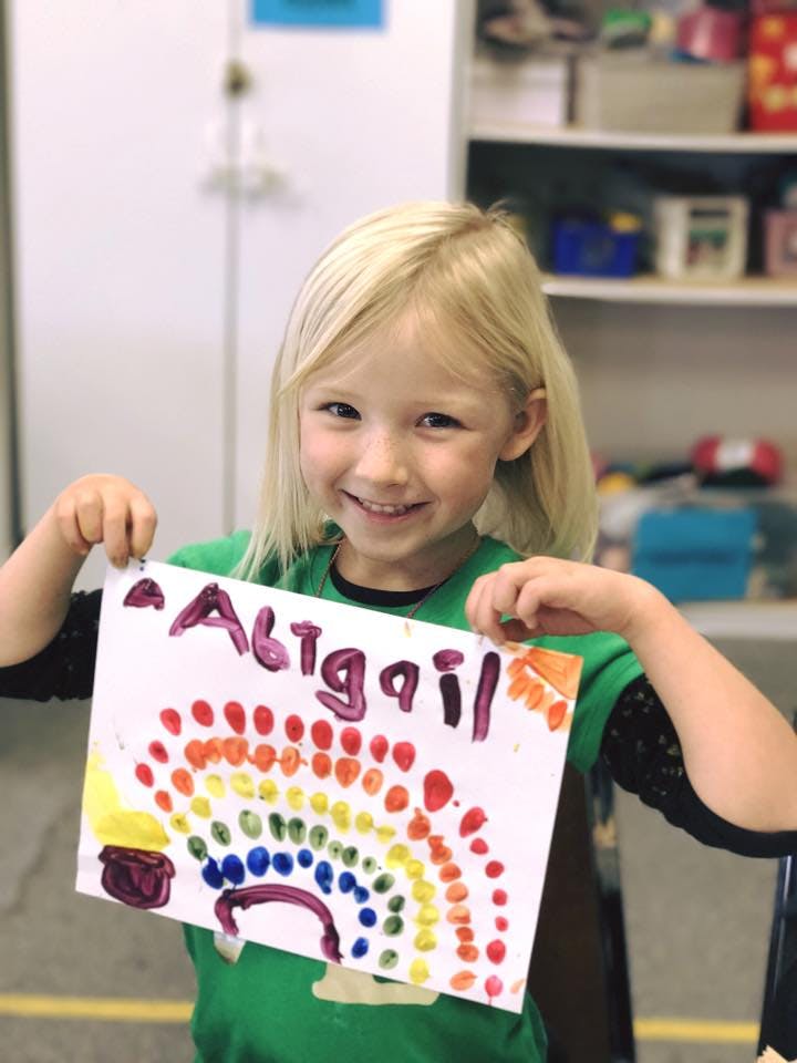 Child holding up a drawing of a rainbow and the name "Abigail"