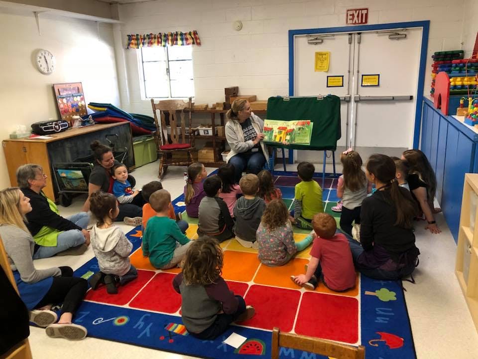 Adult reading a book to a group of children and adults in a classroom