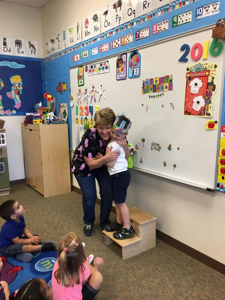 Adult hugging a child standing on a step stool in a classroom