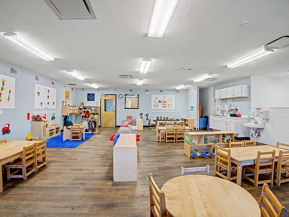 Empty classroom with small wooden tables and chairs, toys, and light blue walls