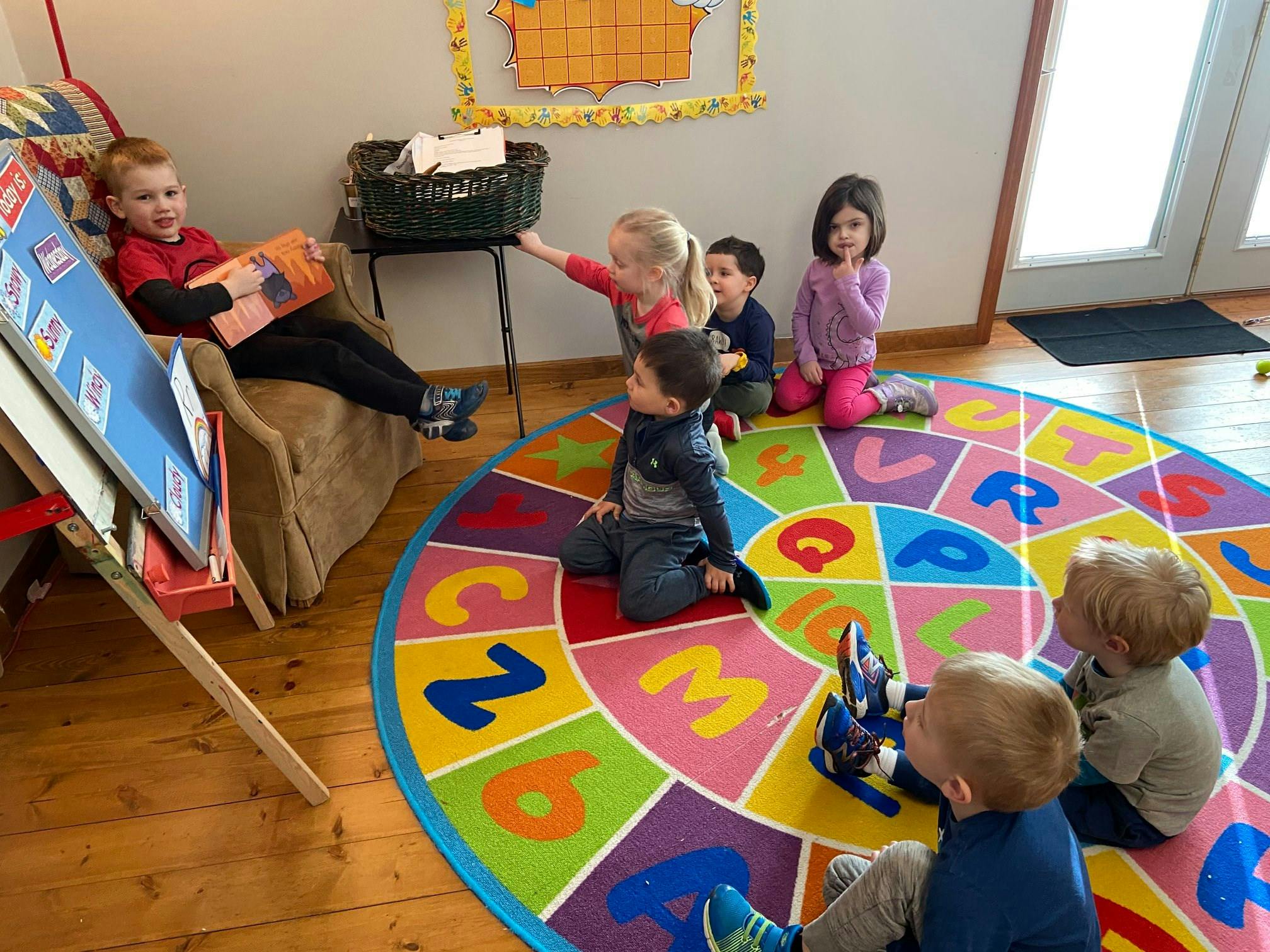 Children sitting on a colorful rug and a child reading a book in a chair