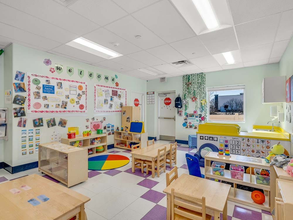Empty classroom with small wooden tables and chairs, toys on shelves, and decorations