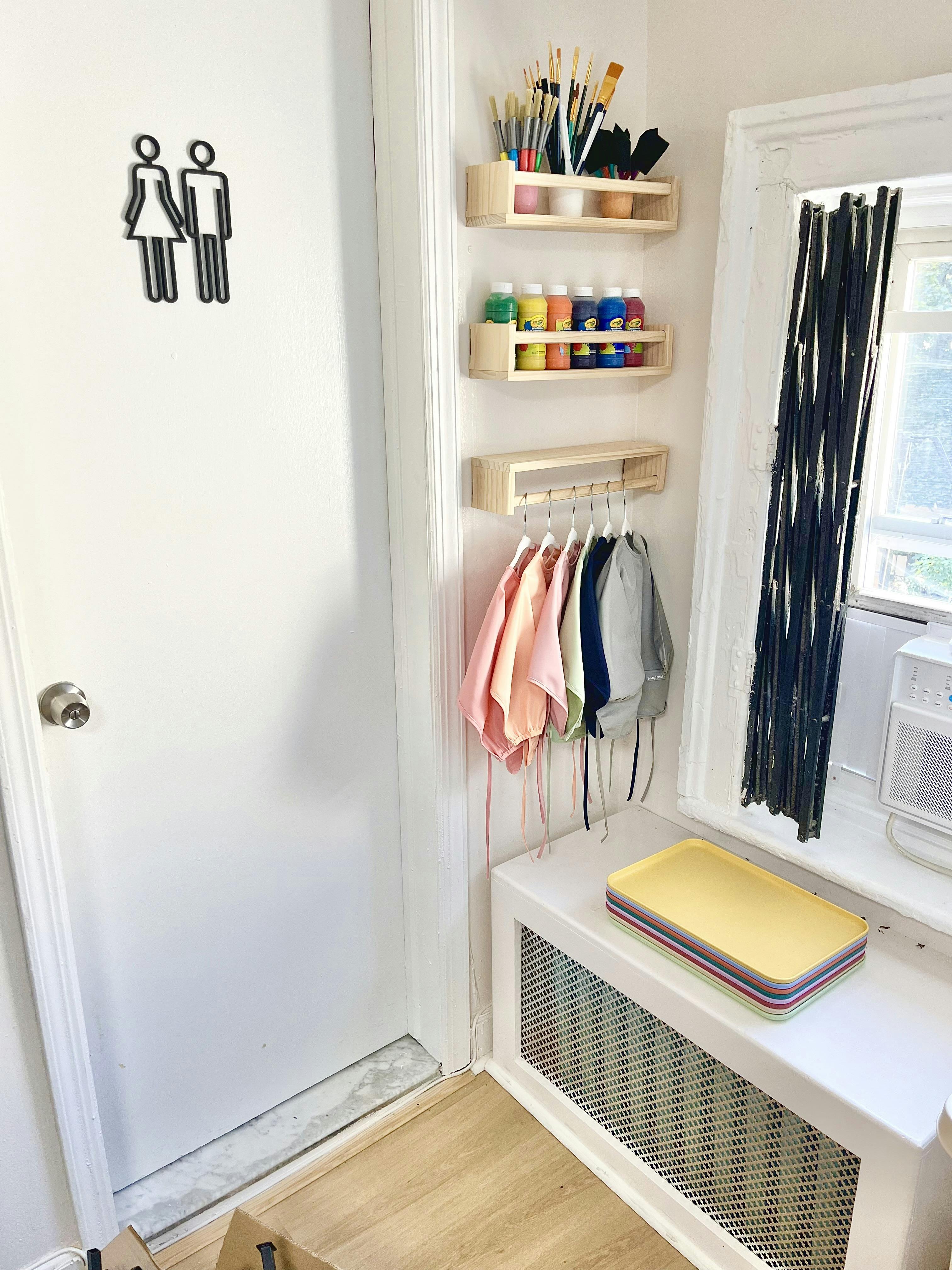 Restroom sign on a white door next to shelves with art supplies and smocks