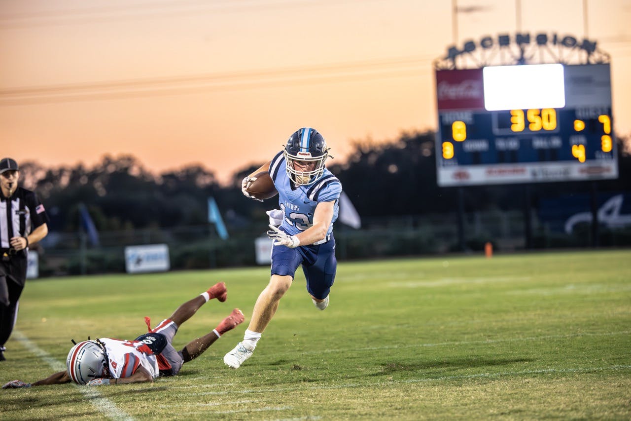 Football player in light blue uniform running with the ball past a fallen opponent