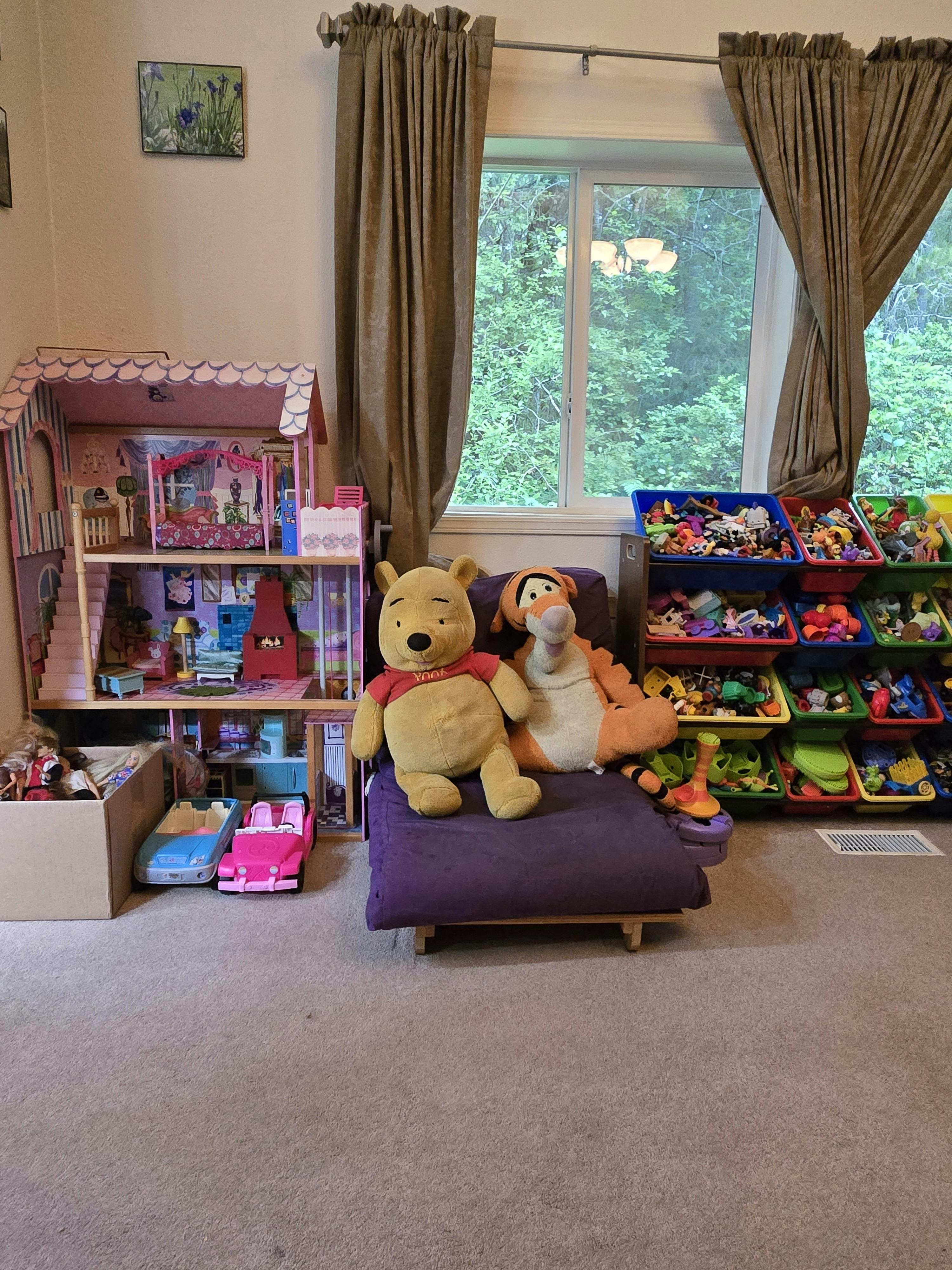 Plush toys sitting on a small chair next to a toy house and bins of toys