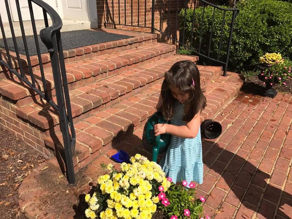 Child holding a watering can near potted flowers on a brick patio
