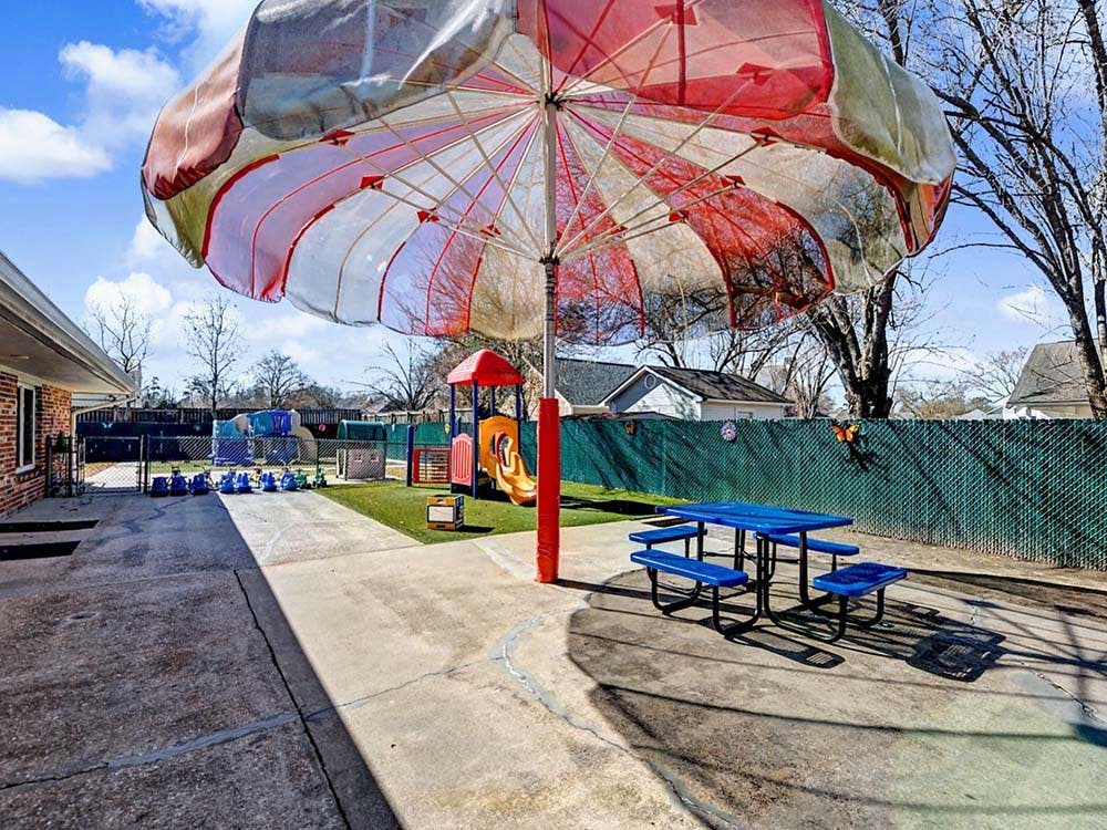 Outdoor playground area with a large umbrella, blue picnic table, and play equipment